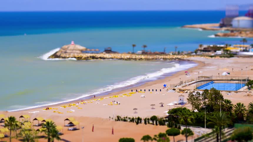 With a tiltshift effect creating a miniaturelike perspective gentle waves roll onto a wide sandy shore where people gather alongside umbrellas and lounge chairs while a breakwater juts into azure waters under a bright sky