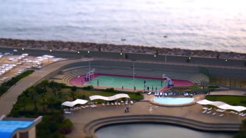 A tiltshift aerial view captures a colorful basketball court with players located next to a swimming pool and the ocean