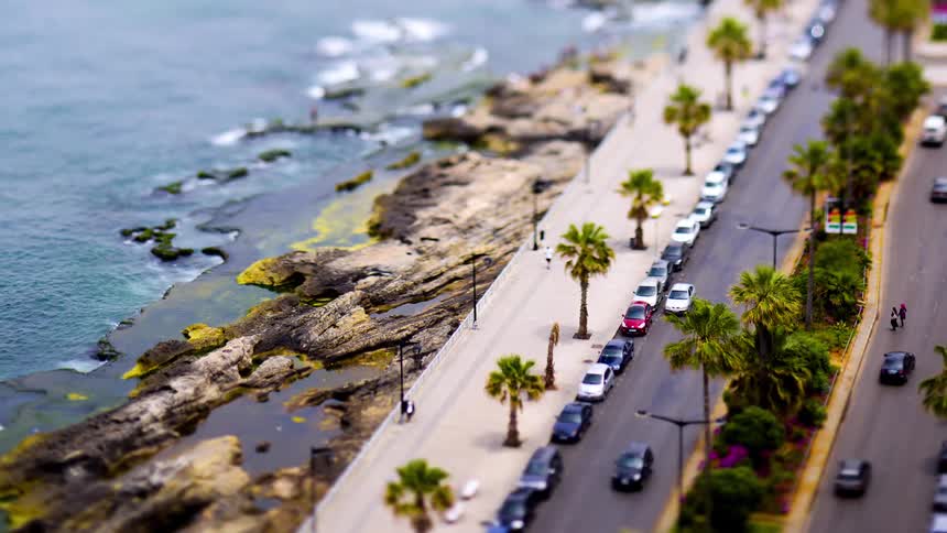 A tiltshift aerial view captures a coastal road lined with palm trees cars and a rocky shoreline