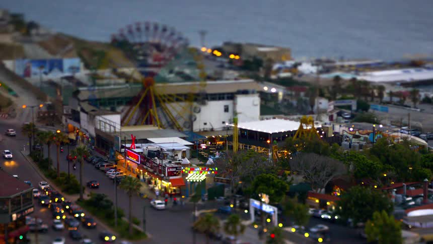 A tiltshift timelapse aerial view shows a bustling city carnival at night with moving traffic a litup ferris wheel blured people and the ocean