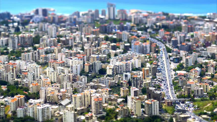 Viewed from above with a tiltshift effect countless buildings winding roads and miniaturelike vehicles appear in a dense cityscape stretching toward the distant coastline under a bright sky capturing a bustling urban environment as cars drive along