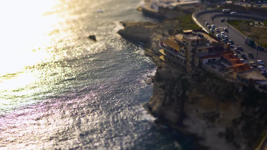 A tiltshift aerial view shows a building perched on a cliff overlooking a sparkling sea and a winding coastal road
