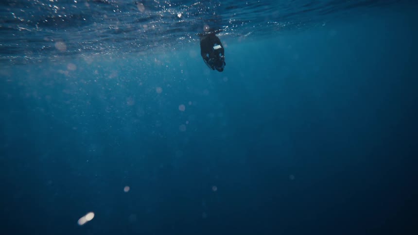 A man floats near the surface underwater with a camera above the deep blue ocean below