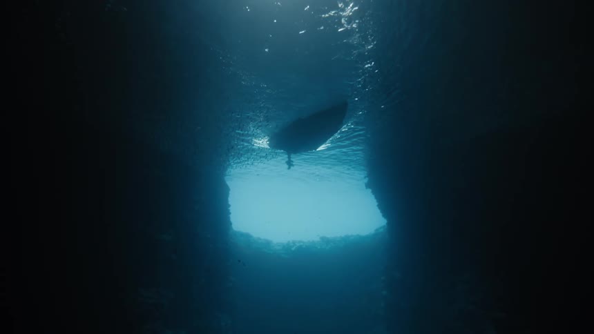 An underwater view shows a motorpropelled boat emerging from a cave surrounded by sunlight filtering through the blue water