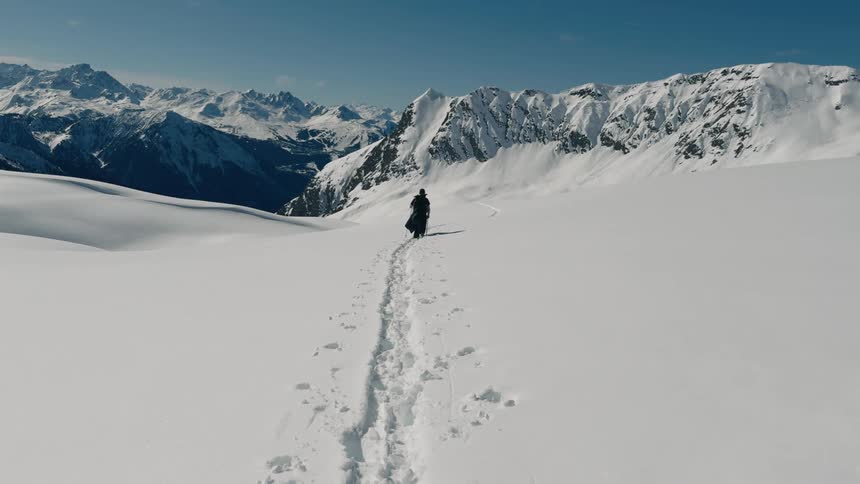 A person is hiking through a vast snowy mountain landscape with snowcovered peaks in the background carrying a large backpack and using trekking poles