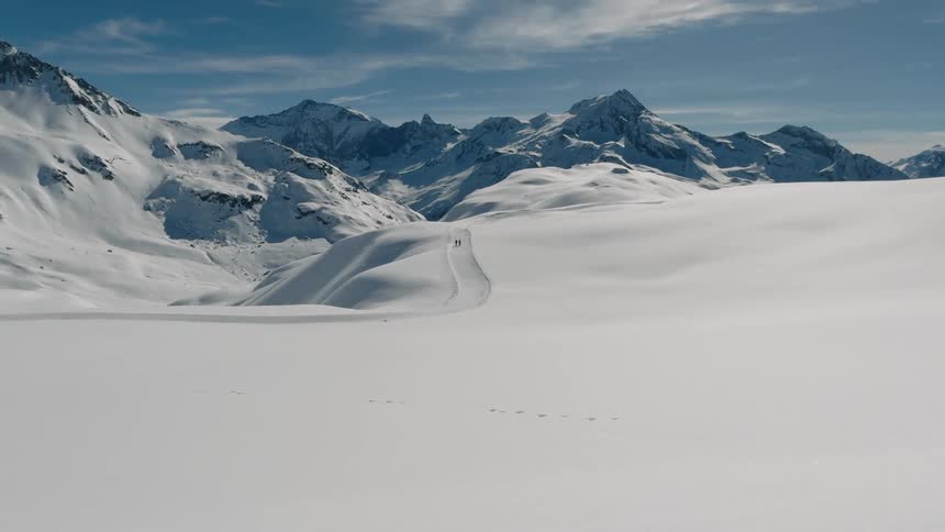 Two distant hikers traverse a vast snow covered landscape leading towards jagged snowcapped mountains under a partially cloudy sky