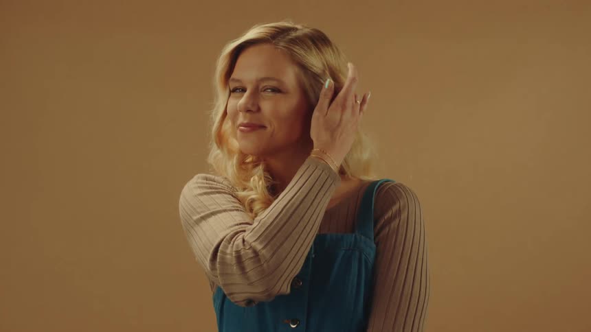 A pretty blonde woman nervously tucks her hair behind her ear as she smiles for the camera