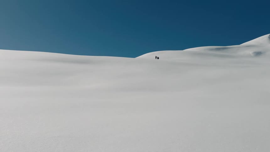 Two small figures walk across a vast snowcovered landscape under a clear blue sky