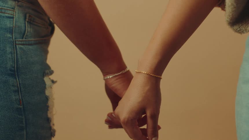 A closeup shows two women holding hands adorned with gold and silver bracelets against a soft beige background