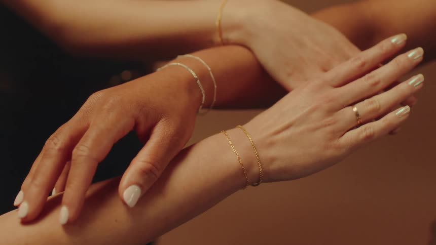 Three arms adorned with delicate gold chain bracelets are intertwined against a softly lit background