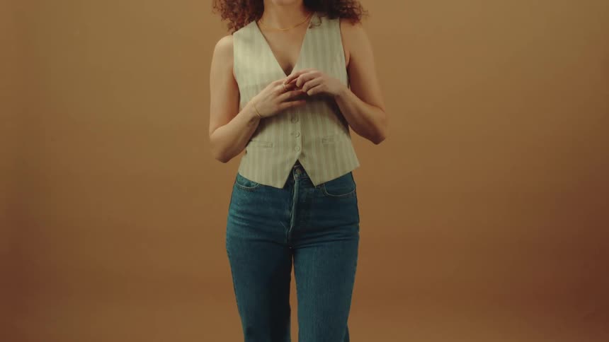 A woman with curly hair dressed in a vest and jeans poses confidently for the camera against a beige background