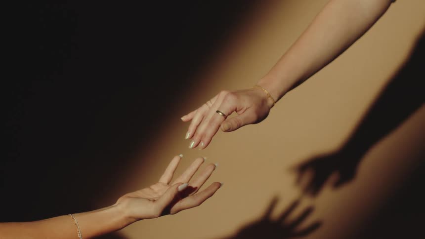 Two hands adorned with ring and bracelets reach toward each other casting soft shadows on a beige background