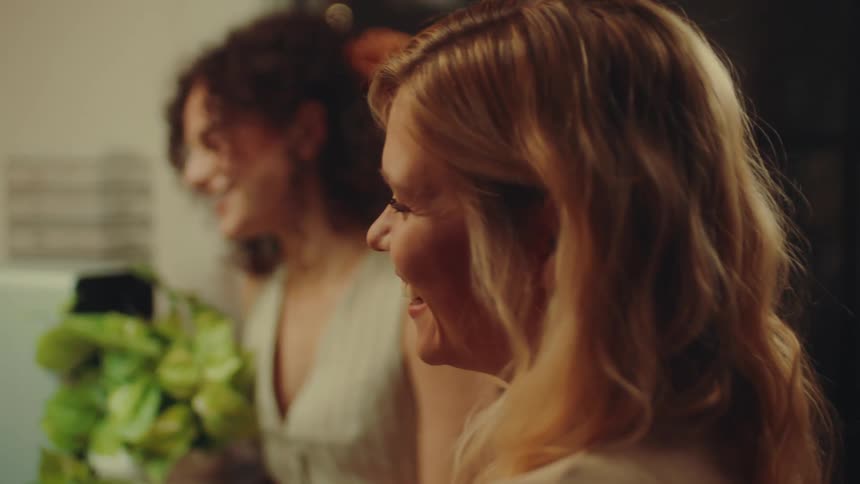 Two women standing near a window at night smile while talking to someone sitting down