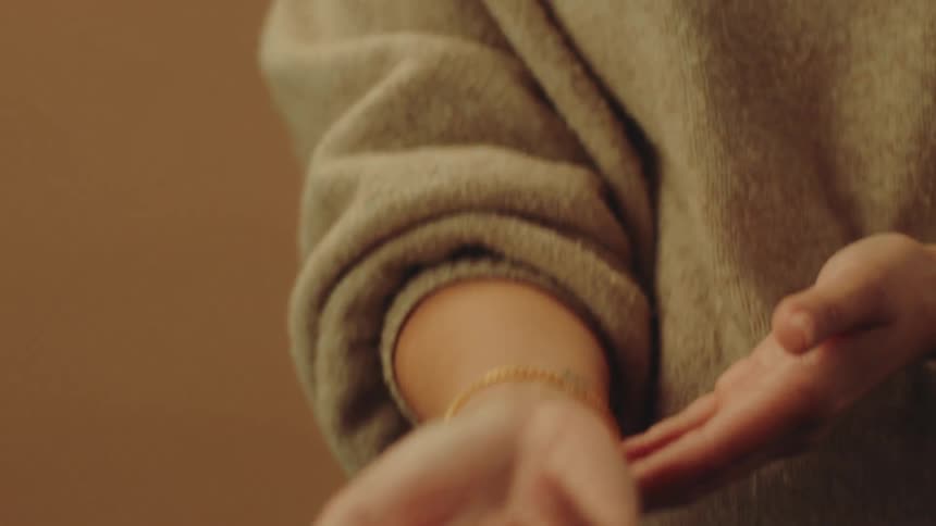 A woman in a sweater adjusts her gold bracelet while looking down and smiling