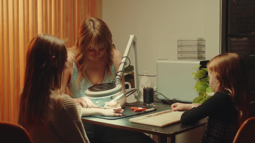 A woman performs a manicure on a girl at a table while another girl sits nearby watching attentively
