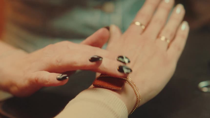 A closeup shows a person positioning a small square brown leather piece beneath a gold chain on another persons wrist