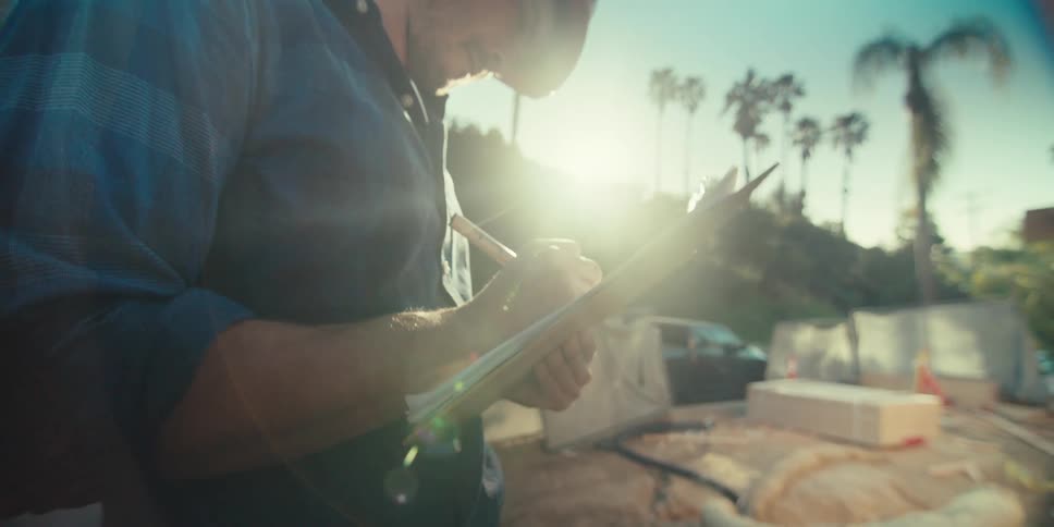 A construction worker wearing a red hardhat takes notes on a clipboard in the morning
