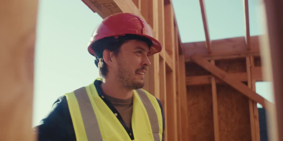 Construction worker hammers in a nail and stands looking around a half built structure