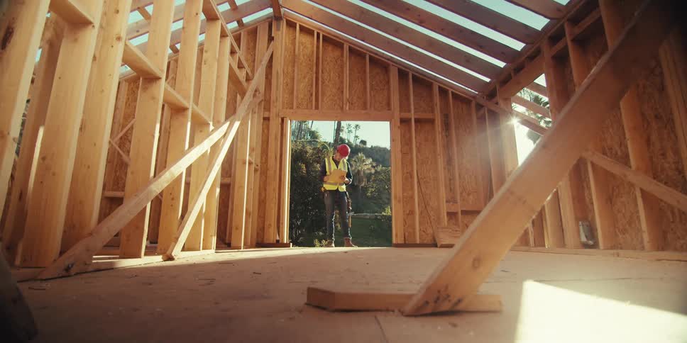 Construction worker stands in the entrance of an unfinished wooden building and makes notes on a clipboard