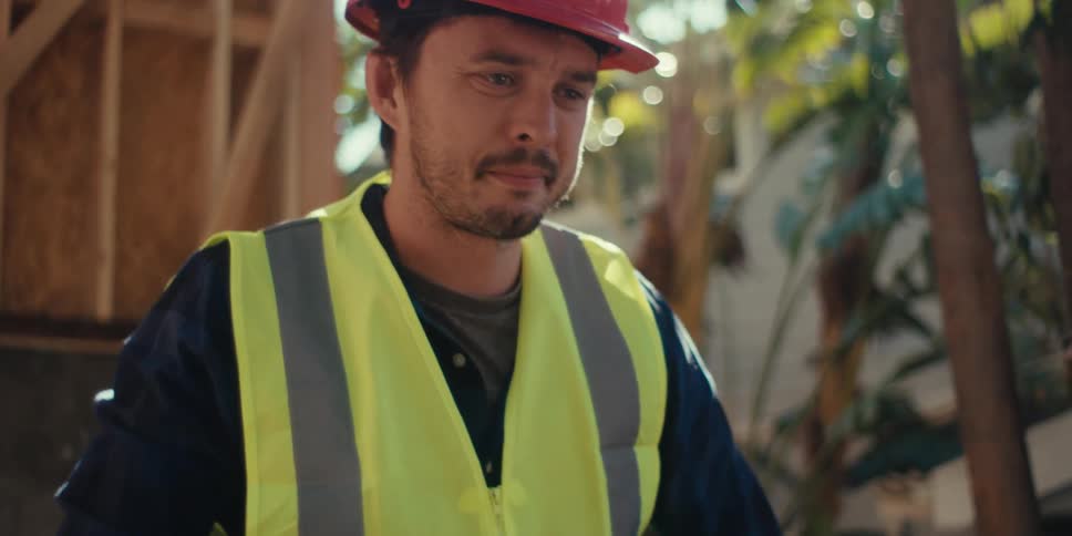 A construction worker wearing a visibility vest and red hard hat uses a level at a construction site