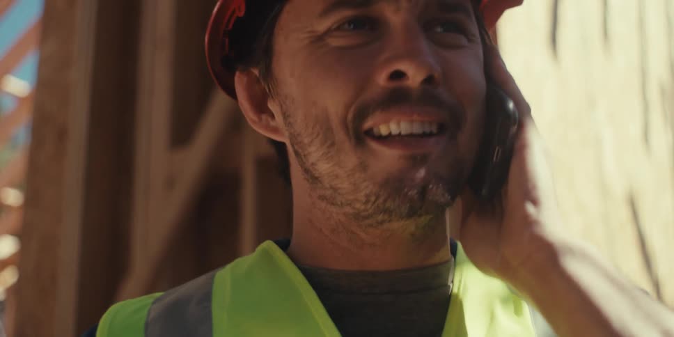 A construction worker wearing a hardhat and a vest makes a phone call