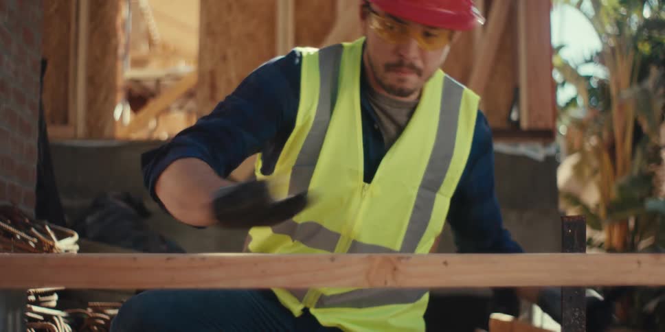 A construction worker wearing a visibility vest and red hard hat uses a level at a construction site