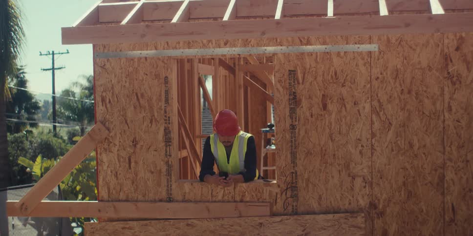 A construction worker in a safety vest and hard hat uses a phone while gazing out through a framed window opening in a partially built wooden structure