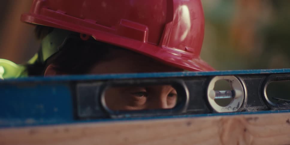 A construction worker wearing a red hardhat checks a level over a beam
