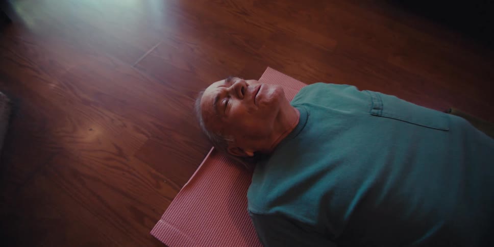 An elderly man in a blue shirt and beige pants is lying on a pink yoga mat on a wooden floor appearing to be resting or meditating in a cozy home setting