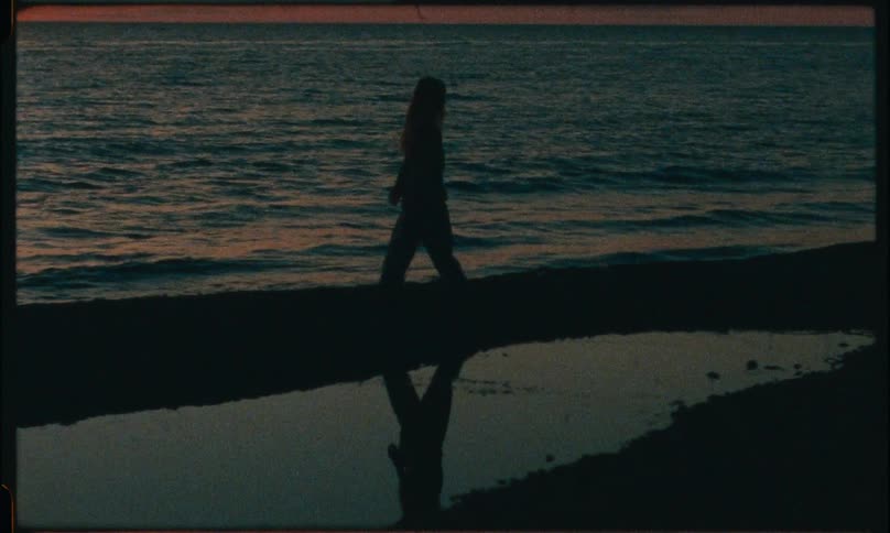 A womans silhouette strolls along a quiet beach at sunset her figure reflected in a small tidal pool as gentle waves roll ashore creating a tranquil dusklit scene