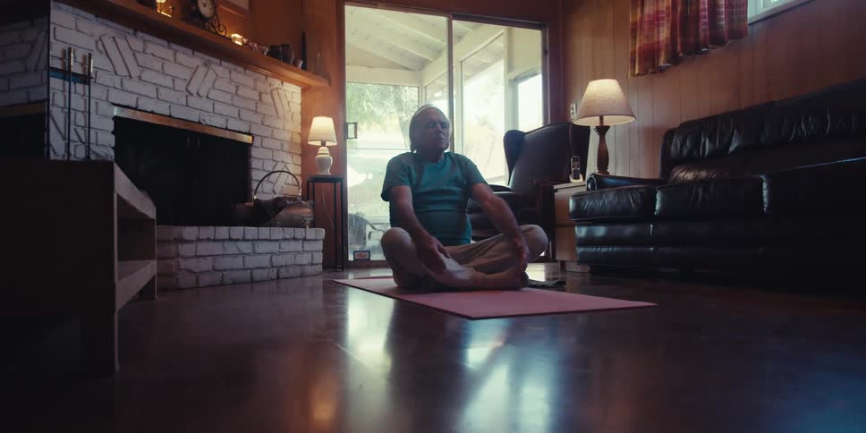 An elderly man is doing a yoga pose on a floor mat indoors