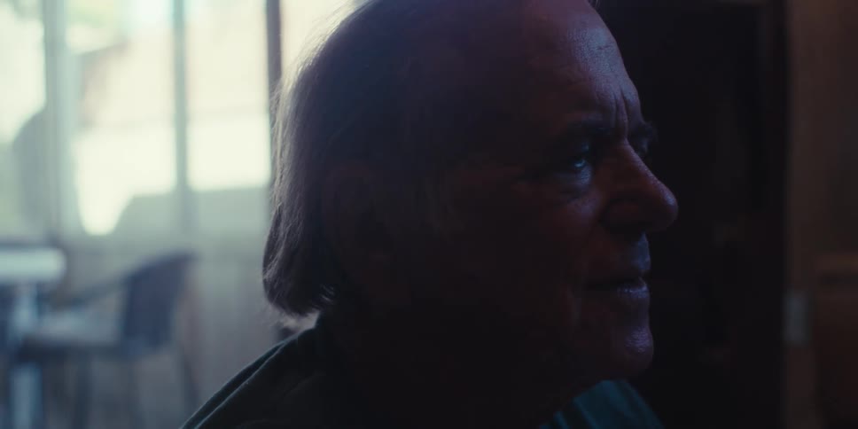 An elderly man sits quietly a gentle smile playing on his lips as he lets out a soft warm laugh framed by the muted light of a window