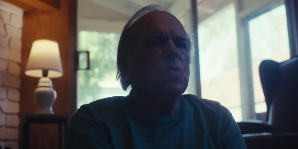 An elderly man sits on the living room floor practicing breathing exercises in a dimly lit room with sunlight streaming through the windows