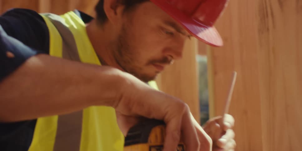 A construction worker wearing a hardhat and a vest takes measurements with a tape