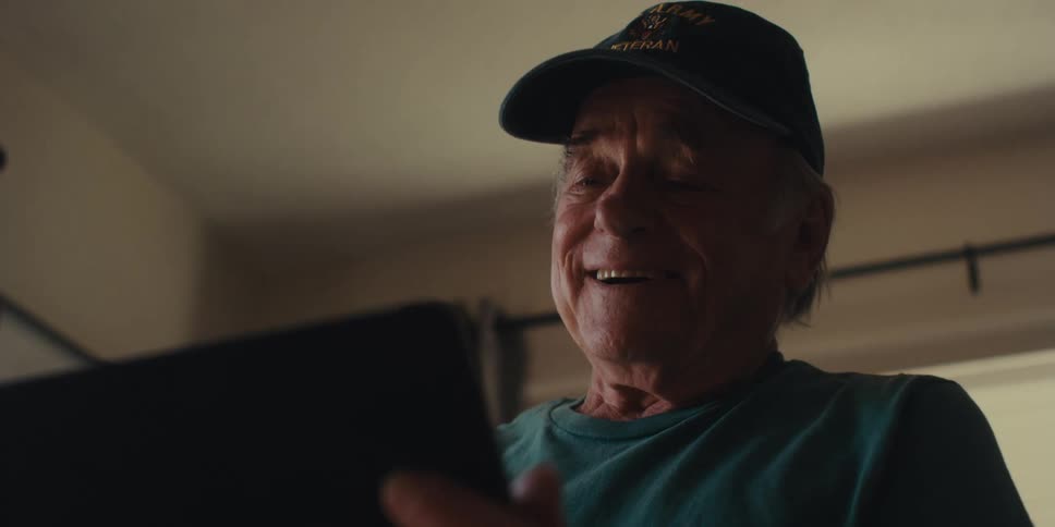 An elderly man in a baseball cap smiles warmly as he looks at a tablet in a cozy home setting