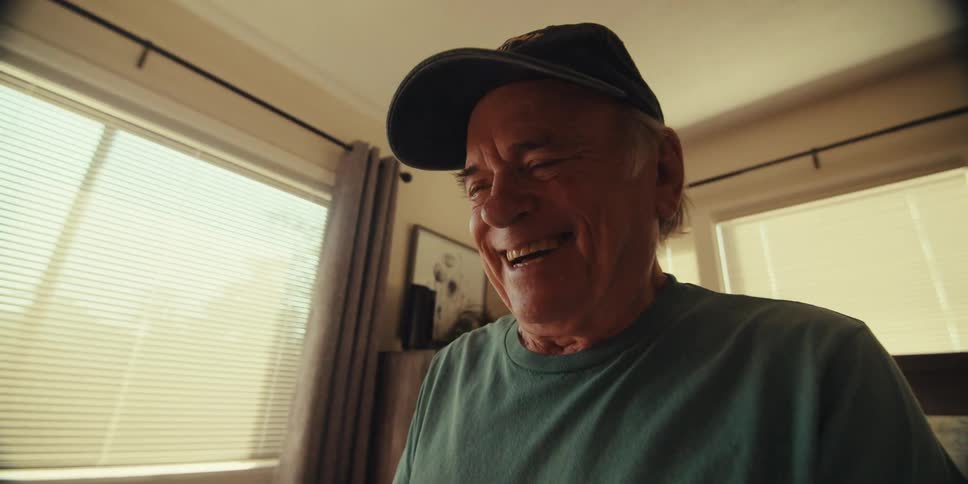 An elderly man wearing a black veteran cap and a blue tshirt sits on a bed looking down at a tablet and laughing in the bedroom