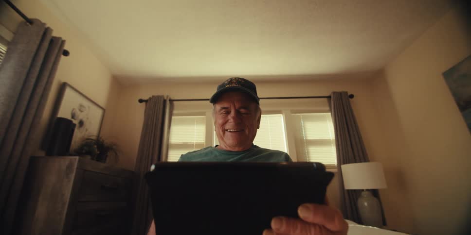 An elderly man in a baseball cap smiles warmly as he looks at a tablet in a cozy home setting