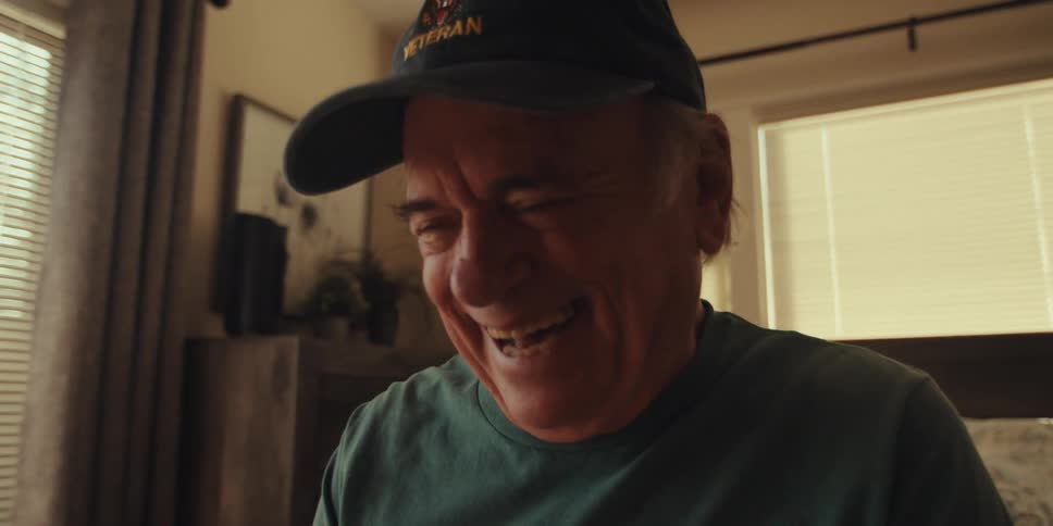 An elderly man sits on a bed in his bedroom laughing while looking at a tablet