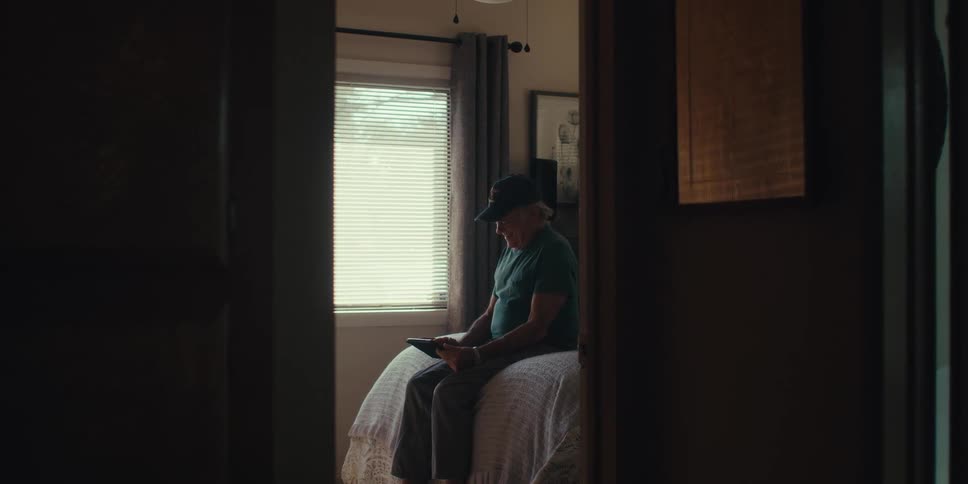 An elderly man sits on a bed in a dimly lit room looking at a tablet with a window behind them and framed pictures on the wall