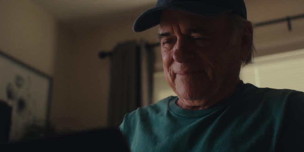 An elderly man sits in a living room laughing at something on a laptop