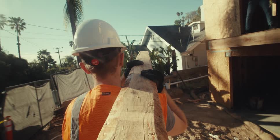 The white man is carrying a long board across the dirt lot to the unfinished house where a black man is waiting in a second story window to pull the board up