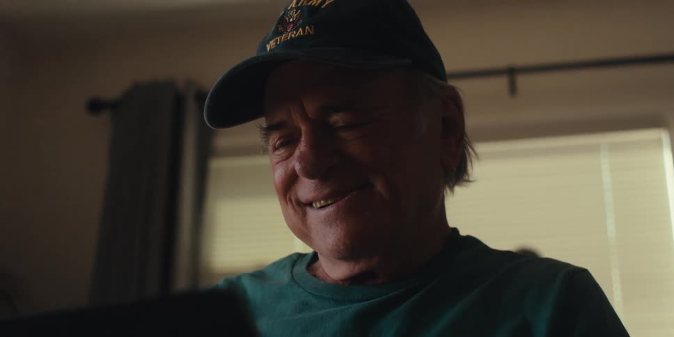 An elderly man in a baseball hat smiles while looking at a tablet in a dimly lit room at home