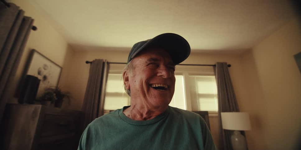 Elderly man sitting on his bed laughs while using his tablet during the day