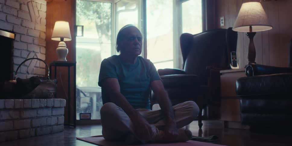 An elderly man sits on the living room floor practicing breathing exercises in a dimly lit room with sunlight streaming through the windows