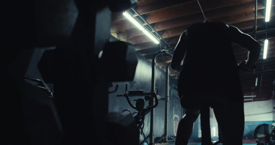 A person exercises intensely on a stationary bike in a dimly lit gym with various workout equipment in the background