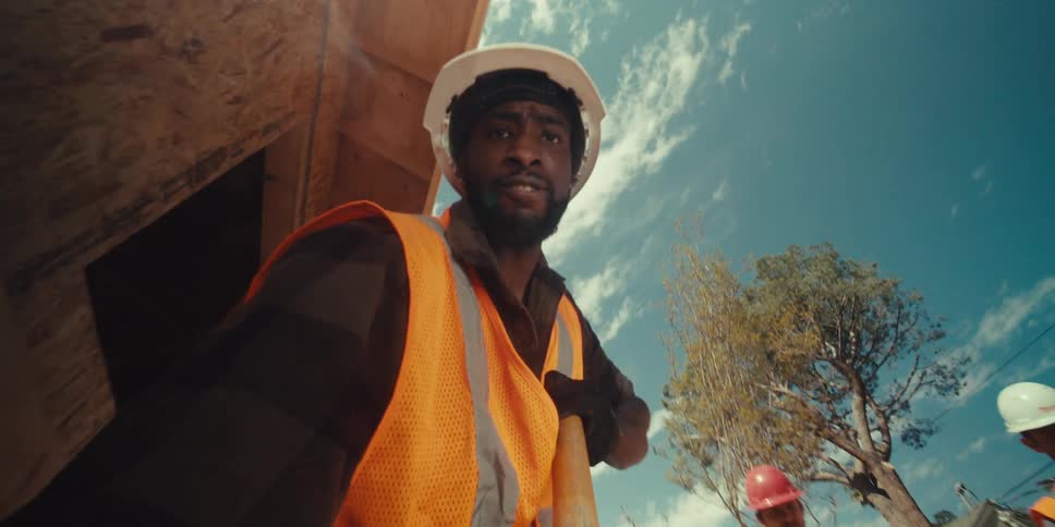 A construction worker wearing a hard hat and safety vest stands at a building site holding a shovel with a blue sky overhead