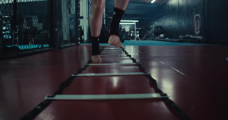 A woman performs agility ladder drills in a dimly lit gym surrounded by various exercise equipment
