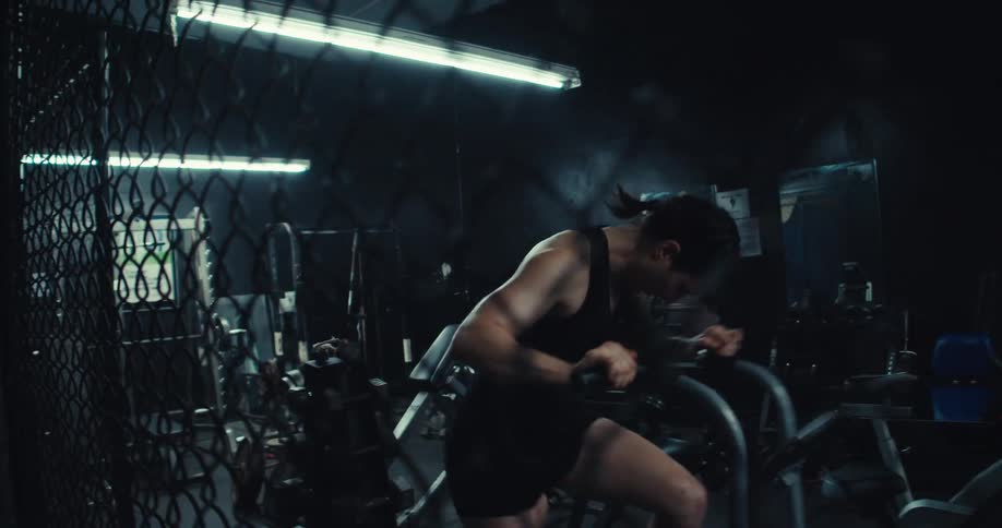 A person exercises intensely on a stationary bike in a dimly lit gym with various workout equipment in the background