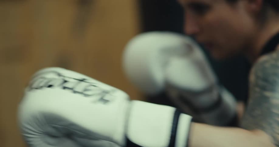 A strong woman with tattooed arms and braided hair practices punching a bag in a gym while wearing white boxing gloves