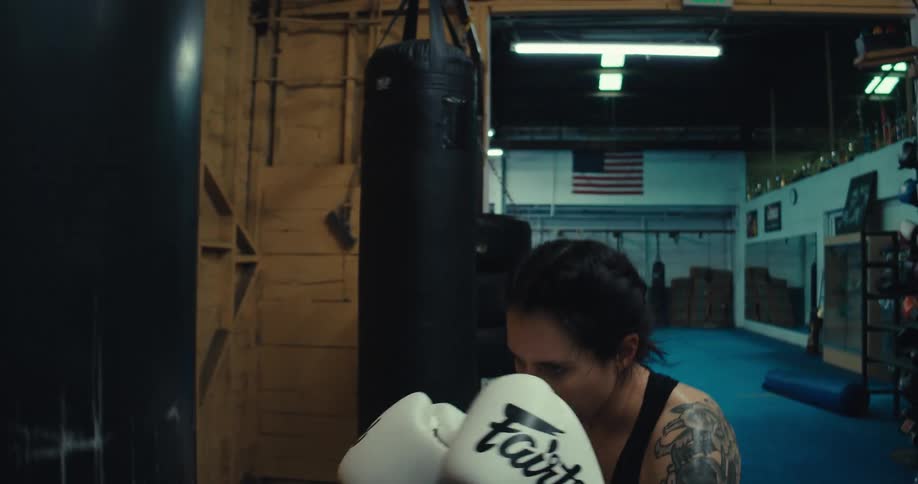 The tattood woman is spinning elbowing and punching the punching bag that is in front of her